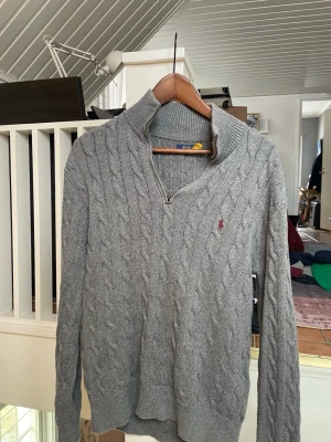 Grå kabelstickad tröja Polo Ralph Lauren - Snygg grå kabelstickad tröja från Polo Ralph Lauren med halv dragkedja och klassisk röd logga på bröstet. Tröjan har hög krage och ribbade muddar vid ärmslut och nederkant. Perfekt för lager på lager och en stilren vardagslook. Defekter på bild 4.                Byten kan vara intressant!