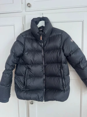 Svart dunjacka från Colmar - Snygg svart dunjacka från Colmar med hög krage och två dragkedjeförsedda fickor. Jackan har ett puffigt quiltat mönster och Colmar-logga på ärmen. Perfekt för kalla vinterdagar och riktigt stilren look.