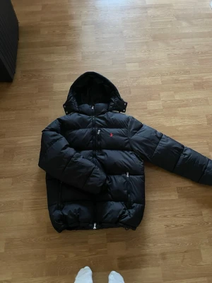 Svart Ralph Lauren pufferjacket - Säljer en svart pufferjacka med huva och dragkedjor framtill. Jackan har en liten röd logga på bröstet och två sidofickor med dragkedja. Perfekt för kalla vinterdagar och riktigt skön att bära. Klassisk, stilren design som passar till det mesta.