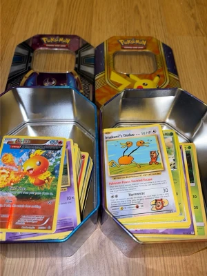 Pokémonkort i samlarburkar - Två metallburkar med Pokémonmotiv fyllda med blandade Pokémonkort. Burkarna har färgglada tryck med Pikachu och Solgaleo på locket. Korten är av olika typer och motiv, både vanliga och holografiska. Perfekt för dig som gillar att samla eller spela Pokémon TCG. Finns både trainers, energys och vanliga kort. Vissa kort har defekter. Kort från ungefär 2015-2018