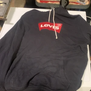 Svart Levi's hoodie med logga - Säljer en svart hoodie från Levi's med stor röd logga på bröstet och vita dragsnören i huvan. Klassisk känguruficka framtill och mjukt material som känns skönt mot huden. Perfekt för dig som vill ha en stilren och bekväm hoodie med streetkänsla.