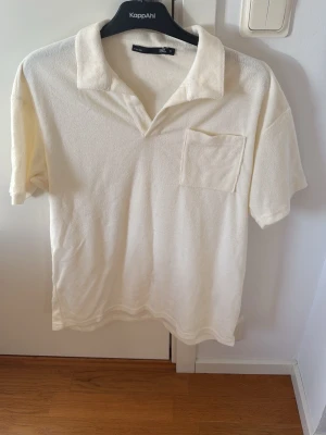 Vit frottétopp från 157 - Vit topp från 157 i mjuk frotté med klassisk krage och bröstficka. Modellen har korta ärmar och en relaxed fit som ger en chill vibe. Perfekt för varma dagar när du vill ha något bekvämt men ändå stilrent.