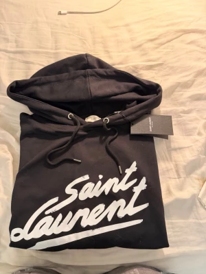 Saint Laurent hoodie  - Snygg svart hoodie från Saint Laurent med stor vit logga i graffiti-stil på bröstet. Hoodien har dragsko i huvan och ribbade muddar. Tillverkad i mjukt bomullsmaterial och har en klassisk streetwear-look. Perfekt för dig som gillar exklusiva märken.