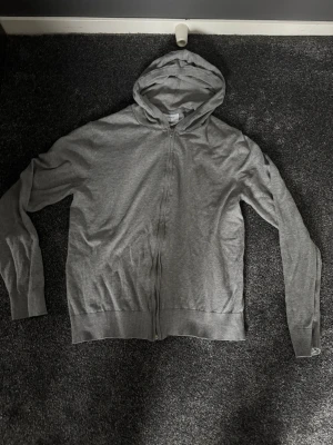 Grå zip hoodie med huva - En enkel och stilren grå hoodie med dragkedja framtill och huva. Tillverkad i mjukt bomullsmaterial som känns skönt mot huden. Perfekt för en avslappnad look och funkar året runt. Klassisk passform och ribbade muddar vid ärmslut och nederkant.