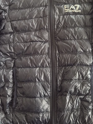 Svart pufferjacka från EA7 Armani - Snygg svart pufferjacka från EA7 Emporio Armani med quiltad design och dragkedja framtill. Jackan har två sidofickor och diskret logga på bröstet. Tillverkad i glansigt syntetmaterial som håller värmen under kalla dagar.