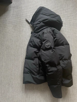Svart dunjacka med huva Canada Goose macmillan - Säljer en svart dunjacka från Canada Goose med stor huva och patch på ärmen. Jackan har en puffig siluett, dragkedja och tryckknappar framtill samt två sidofickor. Perfekt för kalla vinterdagar och riktigt varm tack vare dunfyllningen.