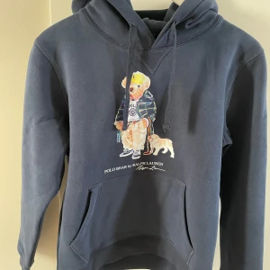 Mörkblå hoodie Polo Bear Ralph Lauren - Mörkblå hoodie från Ralph Lauren med Polo Bear-tryck framtill där björnen har streetwear-outfit och hund. Klassisk känguruficka, dragsko i huvan och mjukt bomullsmaterial. Perfekt för dig som gillar ikoniska prints och avslappnad stil. Otroligt skön! Som ny. (Små i storleken)                                               Inga defekter på tröjan som jag kan se                                                                                Hör av er vid funderingar! 😊