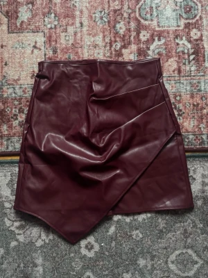 Vinröd mini(ish) kjol i skinn skinnimitation - Säljer en vinröd kort omlottkjol i skinnimitation med asymmetrisk nederkant. Kjolen har en cool drapering framtill och dragkedja nedtill. Är i kortare laget 