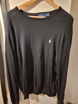 Svart tröja från Polo Ralph Lauren - Klassisk svart tröja från Polo Ralph Lauren med rund halsringning och den ikoniska vita logobroderingen på bröstet. Tillverkad i mjukt material och har ribbade muddar vid ärmslut och nederkant. Perfekt för en stilren look.
