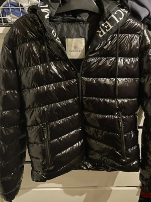 Svart glansig dunjacka från Moncler - Snygg svart dunjacka från Moncler med glansigt material, huva med dragsko och två dragkedjeförsedda fickor. Jackan har Moncler-logga på ärmen och diskret Moncler-tryck innuti huvan. Den har ett litet hål över högra fickan. Pris kan diskuteras! 