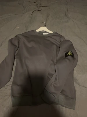 Grå sweatshirt från Stone Island - Snygg grå sweatshirt från Stone Island med klassisk rund hals och ikonisk logotyp-patch på vänster ärm. Tröjan har ribbade muddar vid ärmslut och nederkant, perfekt för en avslappnad och trendig stil.
