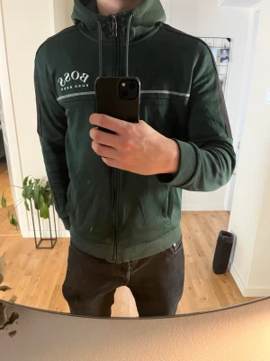 Mörkgrön hoodie från HUGO BOSS - Säljer en mörkgrön hoodie från HUGO BOSS som inte säljs längre, dragkedja framtill och vit logga på bröstet. Väldigt snygg till mörka outfits och till ljusa sommaroutfits.