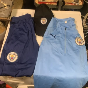 Manchester City träningsset Puma - Snyggt Manchester City träningsset från Puma med ljusblå ziptröja, mörkblå byxor och svart keps. Tröjan har klubbmärke och Puma-logga på bröstet, byxorna har klubbmärke och Puma. Tror ej dom är äkta. Men bra kvalité ändå! Oanvänt.