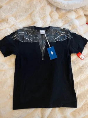 Svart t-shirt med vingmönster från Marcelo Burlon - Säljer en svart t-shirt från Marcelo Burlon med ett detaljerat vingmönster i grått och vitt över axlar och bröst. T-shirten har rund hals och korta ärmar, perfekt för en edgy streetwear-look.