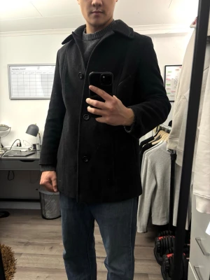 Svart ullrock från LEBEK - Klassisk svart ullrock från LEBEK med tre knappar framtill och bred krage. Rocken har raka ärmar och två sidofickor. Perfekt för kyliga dagar och ger en stilren look till alla outfits.