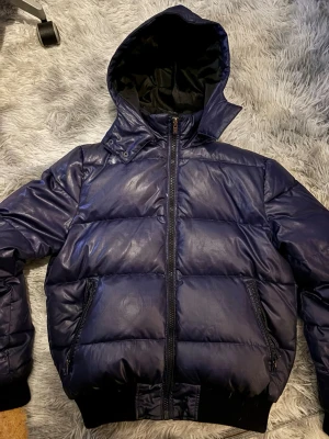 Mörkblå pufferjacka med huva - Säljer en mörkblå / lila pufferjacka med stor huva och dragkedja framtill. Är självklart öppen för prisförslag och säljer fler varor så köper du många får du också mängdrabatt. Obs säljes som på bild.