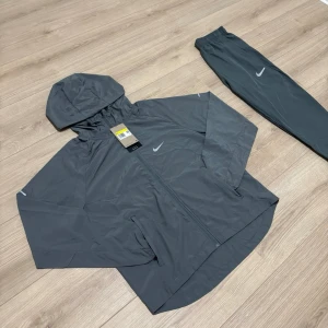 🩶Grå nike repel miller med huva🩶 - Snyggt gråt träningsset från Nike med jacka och byxor. Jackan har huva, dragkedja och vit Nike-logga på bröstet. Byxorna har resår i midjan och vid benslut samt vit Nike-logga på låret. Perfekt för träning eller chill. Tar emot bud. OBS! Den är grå.