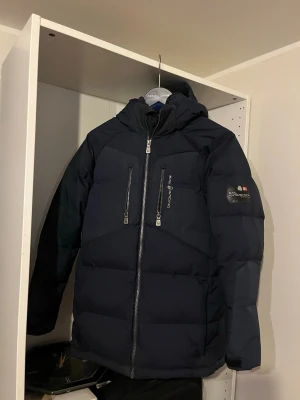 Sail Racing jacka barn 170 - Snygg mörkblå pufferjacka från sail racing med två vertikala bröstfickor samt inneficka med dragkedja, huva och patch på ärmen. Storlek 170, nypris kostar 2600kr på kidsbrandstore.