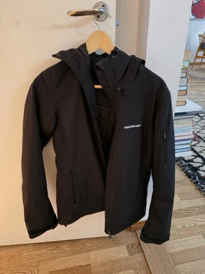  Peak Performance skidjacka  - Snygg svart skidjacka från Peak Performance med huva och dragkedja framtill. Jackan har två sidofickor med dragkedja och en extra ficka på ärmen.