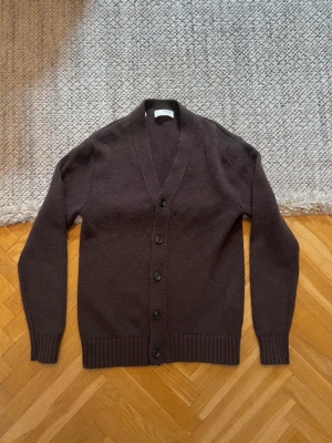 Kofta  - Klassisk mörkbrun kofta med v-ringning och fem knappar framtill. Koftan har ribbade muddar vid ärmslut och nederkant, samt en mjuk och skön stickad känsla. Perfekt för lager på lager och enkel att matcha med olika outfits.