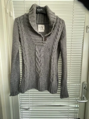 Grå kabelstickad tröja från H&M - Snygg grå kabelstickad tröja från H&M med stor krage och en dekorativ knapp framtill. Tröjan har långa ärmar, ribbade muddar och ett klassiskt flätmönster både fram och på ärmarna. Perfekt för kyliga dagar och enkel att matcha.