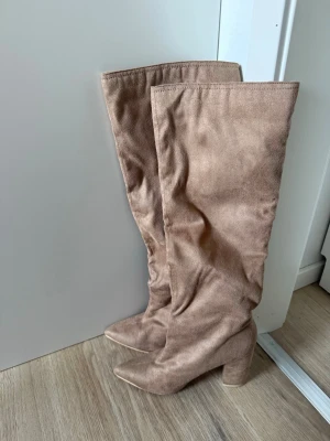 Beige overknee stövlar med klack - Säljer ett par snygga beige overknee stövlar med bred klack och spetsig tå. Stövlarna är i mjukt mockaimitationsmaterial och har en stilren look som passar till många outfits. Perfekta för dig som vill ha ett statement-plagg till hösten eller vintern.