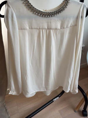 Vit blus med strass från Zara Woman - Stilren vit blus från Zara Woman med långa ärmar och rund halsringning dekorerad med glittrande strassdetaljer. Tillverkad i ett mjukt, lätt material som ger ett snyggt fall. Perfekt för dig som vill ha en elegant touch på din outfit.