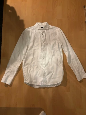 Vit skjorta Riley - Klassisk vit skjorta från Riley med lång ärm och knappar framtill. Skjortan har en tidlös design med normal passform och är tillverkad i ett mjukt bomullstyg. Perfekt för dig som gillar stilrena och enkla plagg.