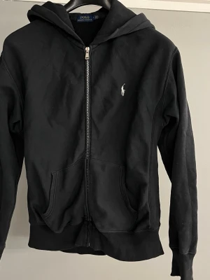 Svart zip hoodie från Polo Ralph Lauren - Klassisk svart hoodie med dragkedja från Polo Ralph Lauren. Tröjan har huva, två fickor framtill och den ikoniska vita logon broderad på bröstet. Perfekt för en avslappnad och stilren look. Storlek M men sitter som en S. Finns några mindre defekter hör av dig för fler bilder. Priset är inte hugget i sten 🪨. 😊👒✌️
