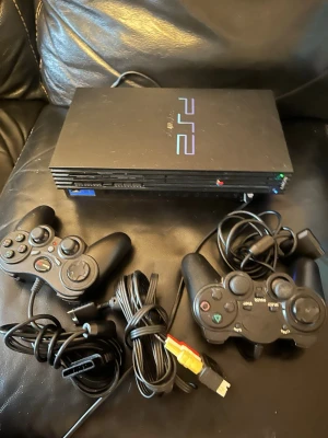Sony PlayStation 2 med två handkontroller - Klassisk svart PlayStation 2-konsol från Sony med två matchande svarta handkontroller och tillhörande kablar. Konsolen har den ikoniska PS2-loggan i blått och lila på ovansidan och är tillverkad i plast med en rektangulär form. Perfekt för retrospel och multiplayer-sessioner.