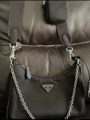 Svart axelväska från Prada med kedja - Snygg svart axelväska från Prada i nylon med silverfärgade detaljer och kedjerem. Väskan har det klassiska Prada-emblemet framtill och en extra liten avtagbar ficka på axelbandet. Perfekt för dig som gillar stilren och trendigt.