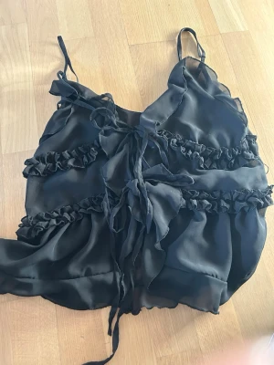 Svart volangig linnetopp med snörning - Svart linnetopp från design by si storlek S. Toppens material är lätt och transparent, vilket ger en luftig känsla. Perfekt för dig som gillar en feminin och trendig look.
