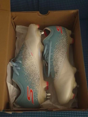 Blåvita fotbollsskor från Skechers Elite  - Snygga fotbollsskor från Skechers i blått och vitt med röda detaljer. Skorna har metall- och plastdobbar för bra grepp på planen och en modern, strömlinjeformad design. Perfekta för dig som vill sticka ut på fotbollsplanen.