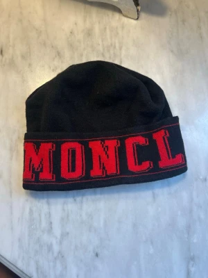 Svart Moncler mössa med röd logga - Säljer en svart stickad mössa från Moncler med bred uppvikt kant och stor röd logotyptext runt hela mössan. Materialet är mjukt och stretchigt, perfekt för kalla dagar. Snygg och enkel att matcha till olika outfits.