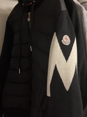 Endast bud Svart Moncler cardigan - Säljer en svart pufferjacka från Moncler med stickade ärmar och ett stort vitt zigzag-mönster på armen. Jackan har huva med dragsko, dragkedja framtill och Moncler-logga på ärmen. Perfekt för dig som vill ha en snygg och varm jacka.