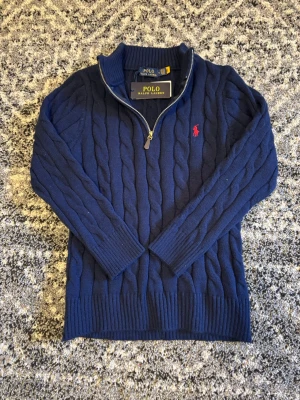 Mörkblå kabelstickad tröja Polo Ralph Lauren - Kabelstickad mörkblå tröja från Polo Ralph Lauren med halv dragkedja och röd broderad logga på bröstet. Tröjan har ribbade muddar och hög krage. Perfekt för lager på lager och klassisk preppy stil. Tröjan är i storlek S. Hör av er vid eventuella frågor eller funderingar!!