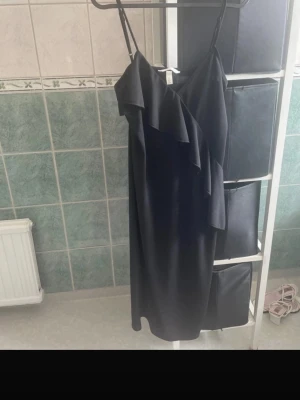 Svart omlottklänning med volang - Snygg svart klänning med tunna axelband och omlottdesign. Klänningen har en volangdetalj framtill som ger en trendig look. Perfekt för dig som gillar stilrena plagg med en twist. Materialet känns mjukt och följsamt.