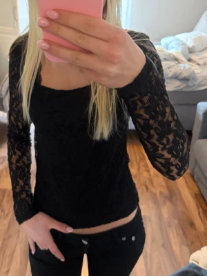 Svart spetsblus med lång ärm - Snygg svart blus i spets med långärmade ärmar och figurnära passform. Blusen har en fyrkantig ringning och är genombruten i materialet vilket ger en trendig look. Perfekt att styla med jeans eller kjol.