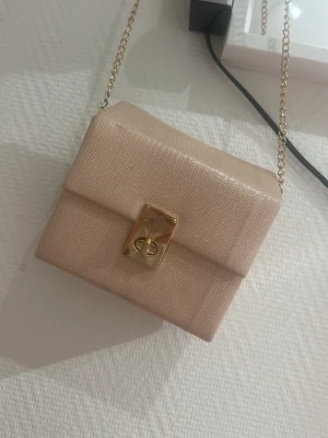 Beige liten axelremsväska med kedja - Snygg beige handväska i boxig modell med guldfärgad kedjerem och elegant låsdetalj framtill. Väskan har en diskret struktur i materialet som ger ett lyxigt intryck. Perfekt för dig som vill ha en stilren accessoar till din outfit.