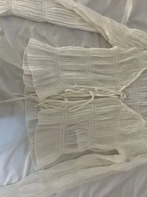 Vit transparent blus med knytband - Trendig vit blus i transparent, plisserat tyg med långa ärmar och dekorativa knytband framtill. Blusen har en luftig och flowy passform som ger en cool och modern vibe. Perfekt att styla lager på lager för en unik look.