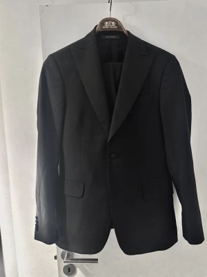 Svart smoking från Oscar Jacobson - Klassiske svart smoking från Oscar Jacobson, modell Elder Blazer. Kavajen har två knappar framtill, spetsiga slag och fickor med lock. Tillverkad i ett elegant tyg som ger en stilren look. Perfekt för dig som vill ha en tidlös och snygg kavaj.