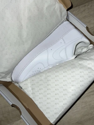 Nike Air Force 1 White - Klassiska helvita Nike Air Force 1 sneakers i läder med låg profil och ikonisk sula. Perfekta för dig som gillar en clean och tidlös stil. Swoosh-logga på sidan och perforerade detaljer vid tån. Helt nya, box lite skadad bara