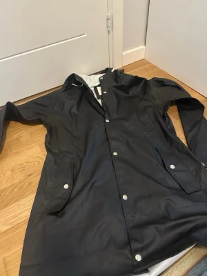 Svart regnjacka med huva stl m - Snygg svart regnjacka med huva och tryckknappar framtill. Jackan har två stora fickor med lock och silverfärgade knappar. Materialet är vattenavvisande och insidan är ljus. Perfekt för regniga dagar.