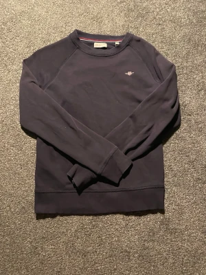 Mörkblå sweatshirt från GANT - Mörkblå sweatshirt från GANT med liten broderad logga på bröstet. Tröjan har rund halsringning med randig detalj på insidan och långa ärmar. Tillverkad i mjuk bomull för en skön och avslappnad stil.