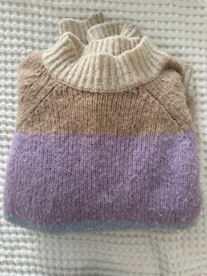 Randig stickad tröja i pastellfärger - Mysig stickad tröja med breda ränder i beige, lila, ljusblått och vitt. Tröjan har en ribbad polokrage samt ribbade muddar vid ärmslut och nederkant. Perfekt för dig som gillar en avslappnad och färgglad stil. Storlek L men passar även mig som har S