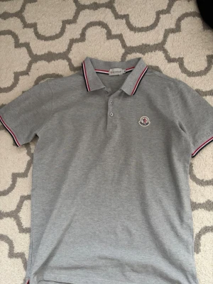 Grå pikétröja från Moncler - Snygg grå pikétröja från Moncler med klassisk krage och knappar framtill. Tröjan har Moncler-logga på bröstet och detaljer i rött och vitt på krage och ärmslut. Perfekt till jeans eller shorts för en clean look.