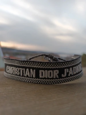 Christian Dior J'Adior tygarmband - Snyggt svart och vitt tygarmband från Christian Dior med texten 'J'Adior' broderat. Armbandet har justerbar knytning och cool, trendig design med kontrasterande färger och detaljerade kanter. Perfekt grisch accessoar för att lyfta din outfit.