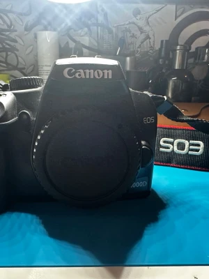 Canon EOS 1000D systemkamera - Canon EOS 1000D är en svart systemkamera i plast med tydliga knappar och stor display på baksidan. Kameran har en klassisk form och levereras med en axelrem märkt EOS. Perfekt för dig som vill ta snygga bilder och utveckla ditt fotograferande. (Lins och batteri saknas)