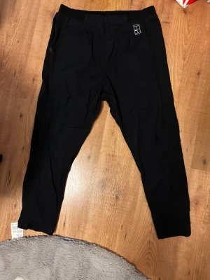 Svarta träningsbyxor från NIKECourt heritage - Svarta mjukisbyxor från NIKE med avslappnad passform och resår i midjan. Byxorna har diskret logga på vänster sida och dragkedjeficka. Perfekta för träning eller chill. Materialet känns mjukt och stretchigt.