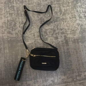 Svart axelremsväska från Pull&Bear - Snygg svart axelremsväska från Pull&Bear med justerbar rem och guldfärgade detaljer. Väskan har en praktisk dragkedja framtill och en dekorativ tag med loggan. Tillverkad i ett slitstarkt tygmaterial och har en stilren, rektangulär form.